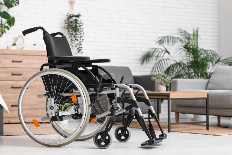 Wheelchair in a living room, near a wooden cabinet, sofa, and plants, with a white brick wall in the background.