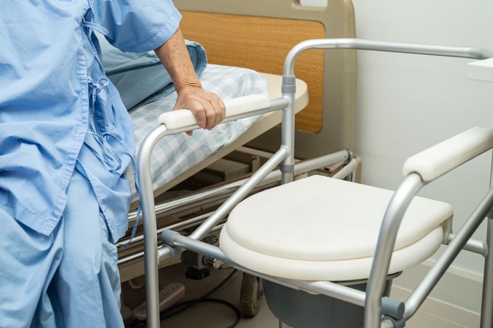 Person in blue hospital gown using a bedside commode next to a hospital bed.