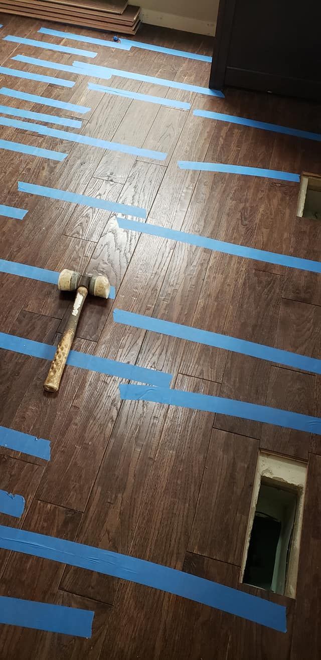 Wood floor marked with blue tape strips, a mallet near the bottom left, and a rectangular cutout in the lower right.