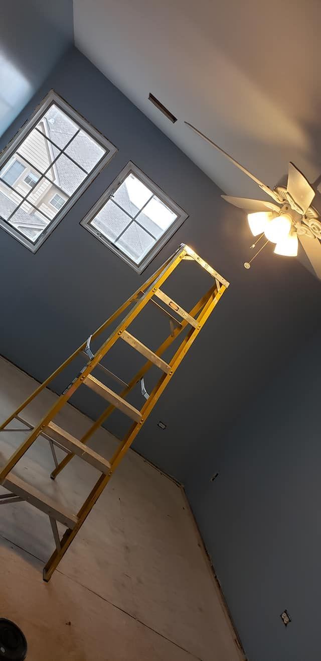 Yellow ladder in a gray room beneath two windows and a ceiling fan.