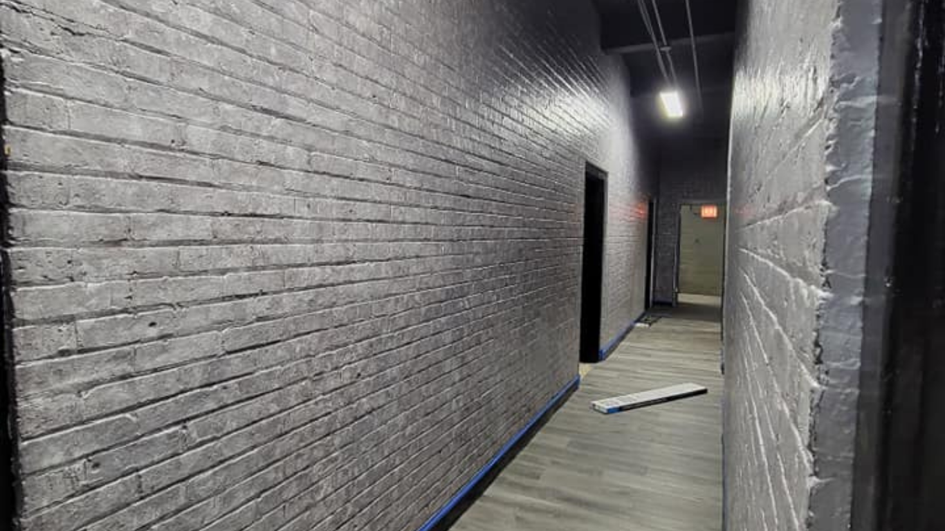 Narrow gray brick hallway with a partially open door and a loose floor panel near the far end.