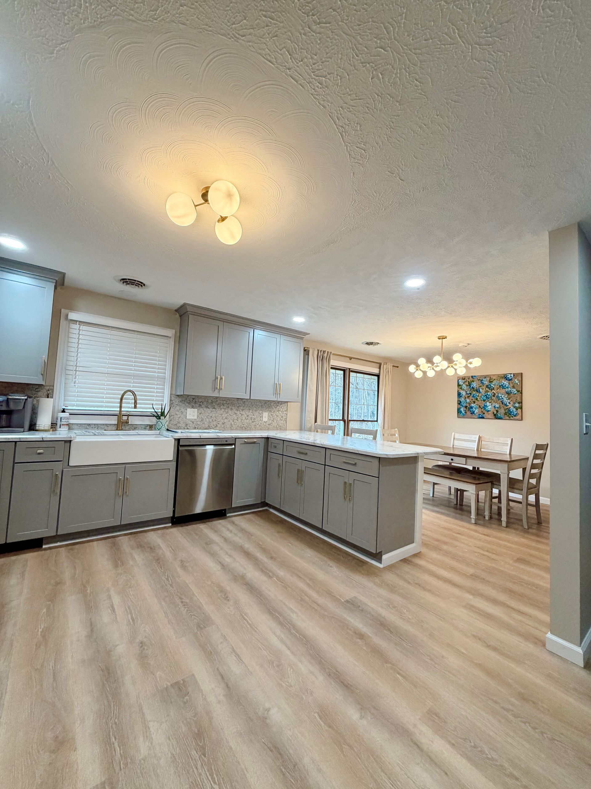 A kitchen with grey cabinets, light wood-look floors, a farmhouse sink, and an adjacent dining area with a chandelier.