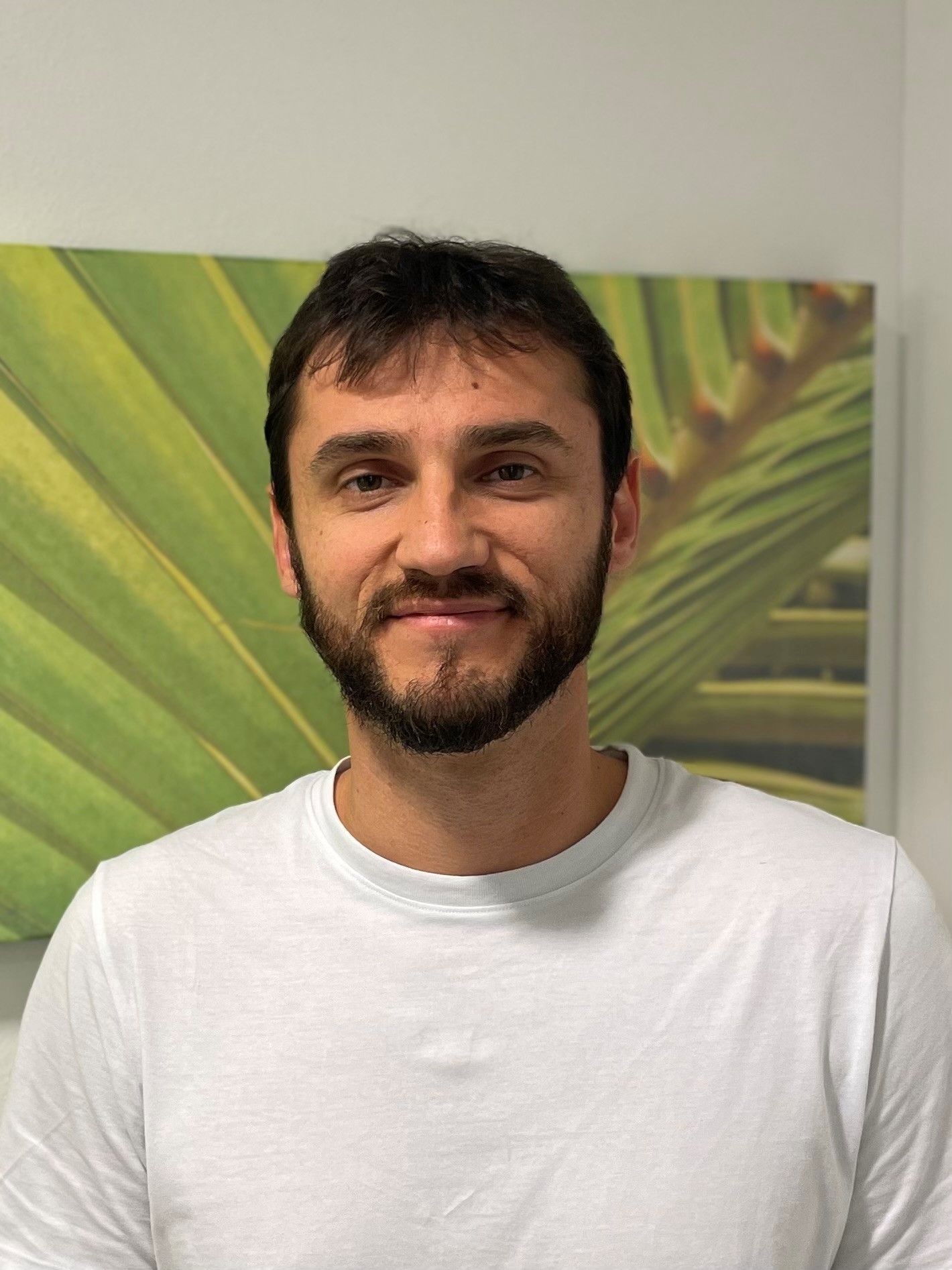 Ein Mann mit Bart und weißem Hemd steht vor einem Gemälde mit Palmblättern.