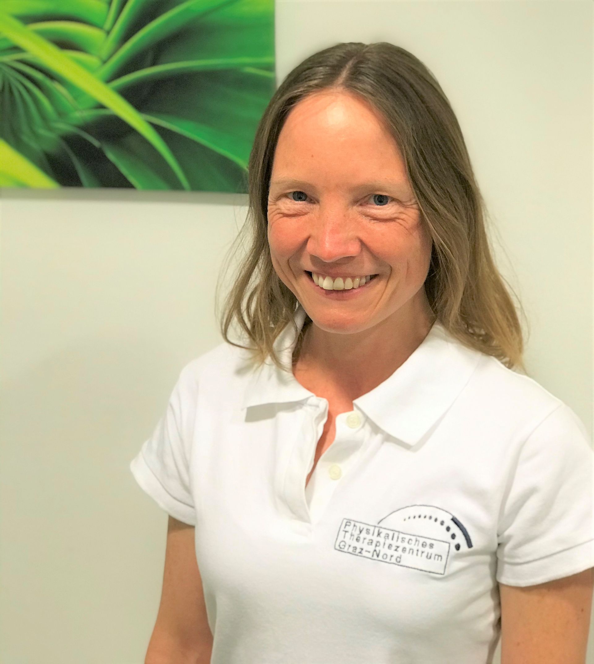 Eine Frau in einem weißen Poloshirt lächelt vor einem Gemälde an der Wand.