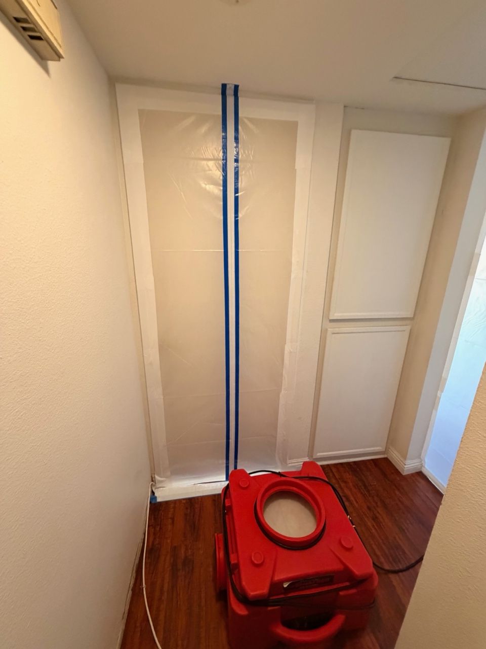 Red dehumidifier in room, plastic sheeting taped to doorway. Wooden floor, white walls.