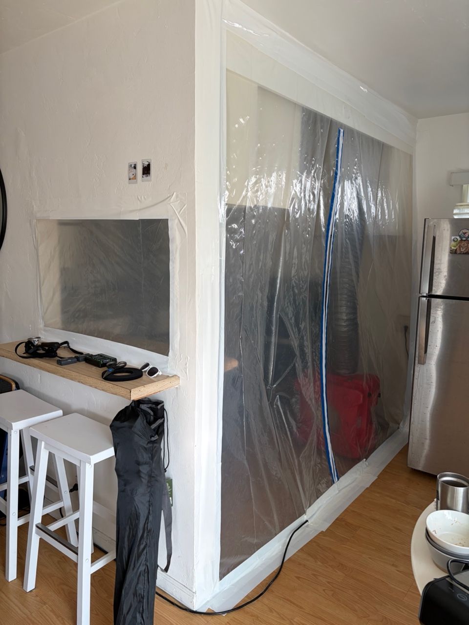 Room corner with plastic sheeting taped to the walls; kitchen with breakfast bar, stools, and stainless steel fridge.