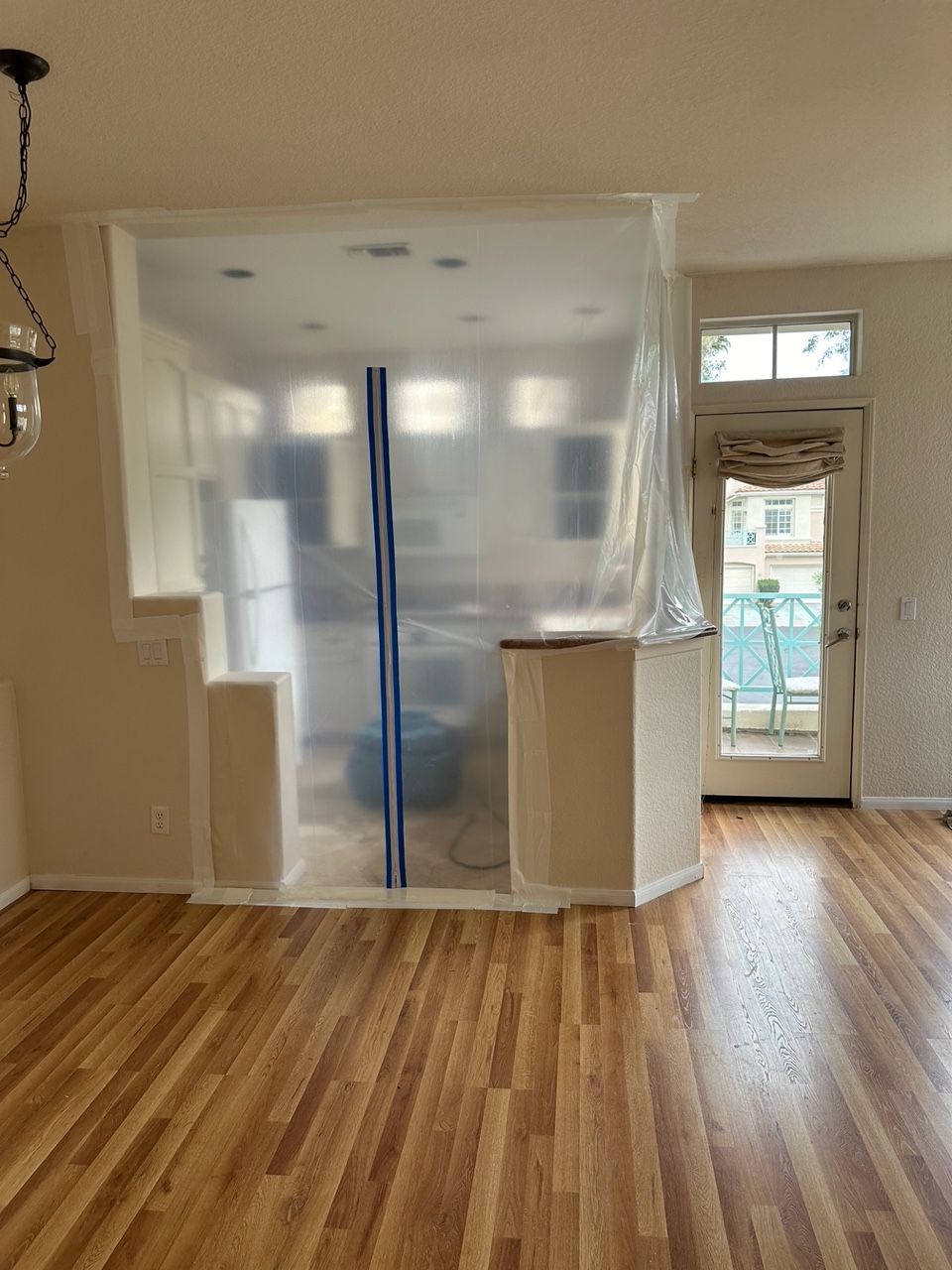 A room with hardwood floors, a kitchen area is sealed off by plastic sheeting. A door leads to a balcony.