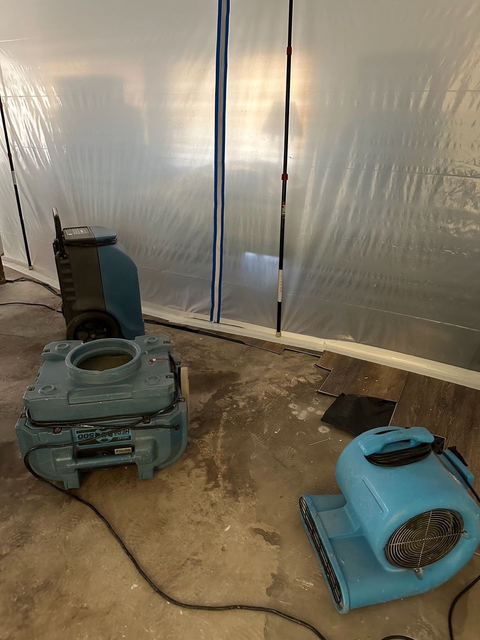 Blue drying equipment inside a plastic-draped area; likely water damage restoration.
