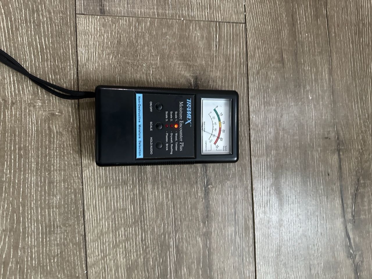 Black EMF meter on wood floor. Meter has a needle gauge, buttons, and a wrist strap.