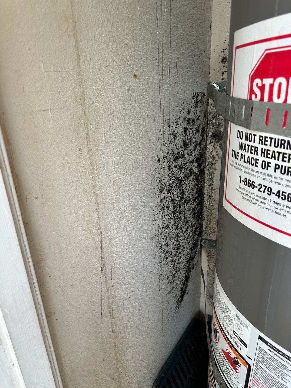 Black splatters on a wall next to a water heater. The heater has a red and white warning label.