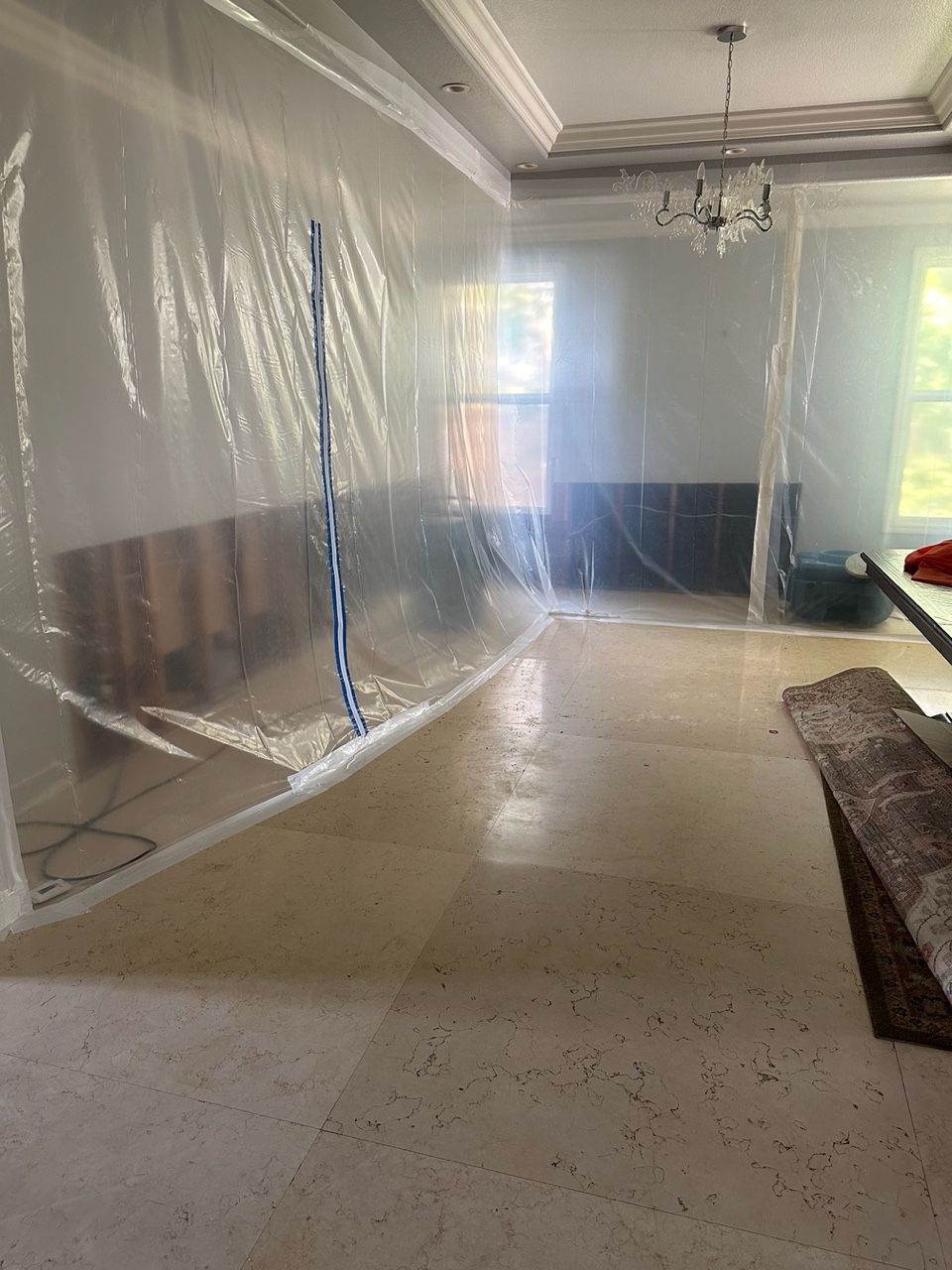Room interior with plastic sheeting covering walls and furnishings, light-colored floor, and chandelier.