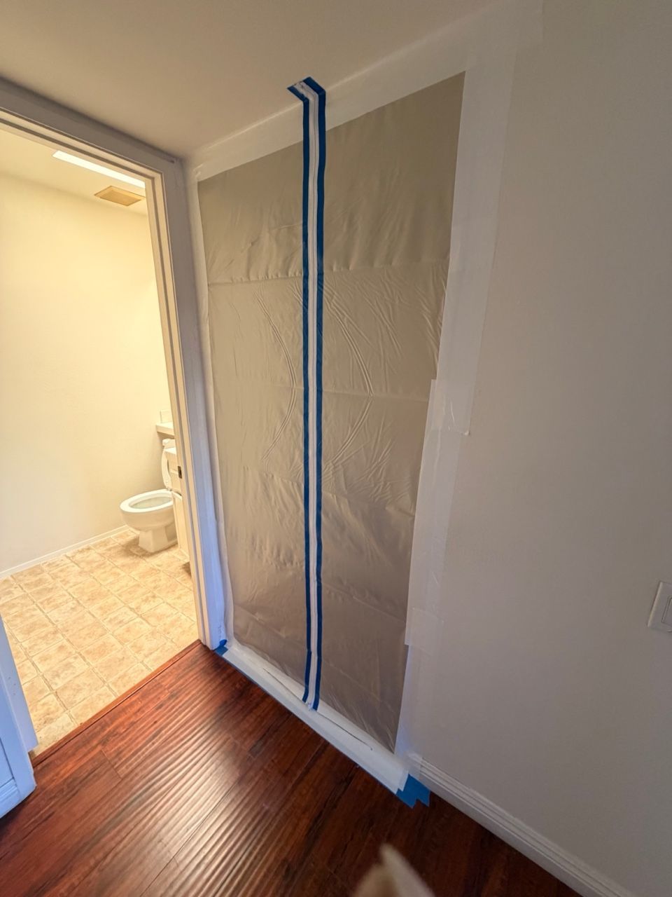 Doorway masked with plastic sheeting, blue painter's tape, leading to a bathroom with a toilet and tan flooring.