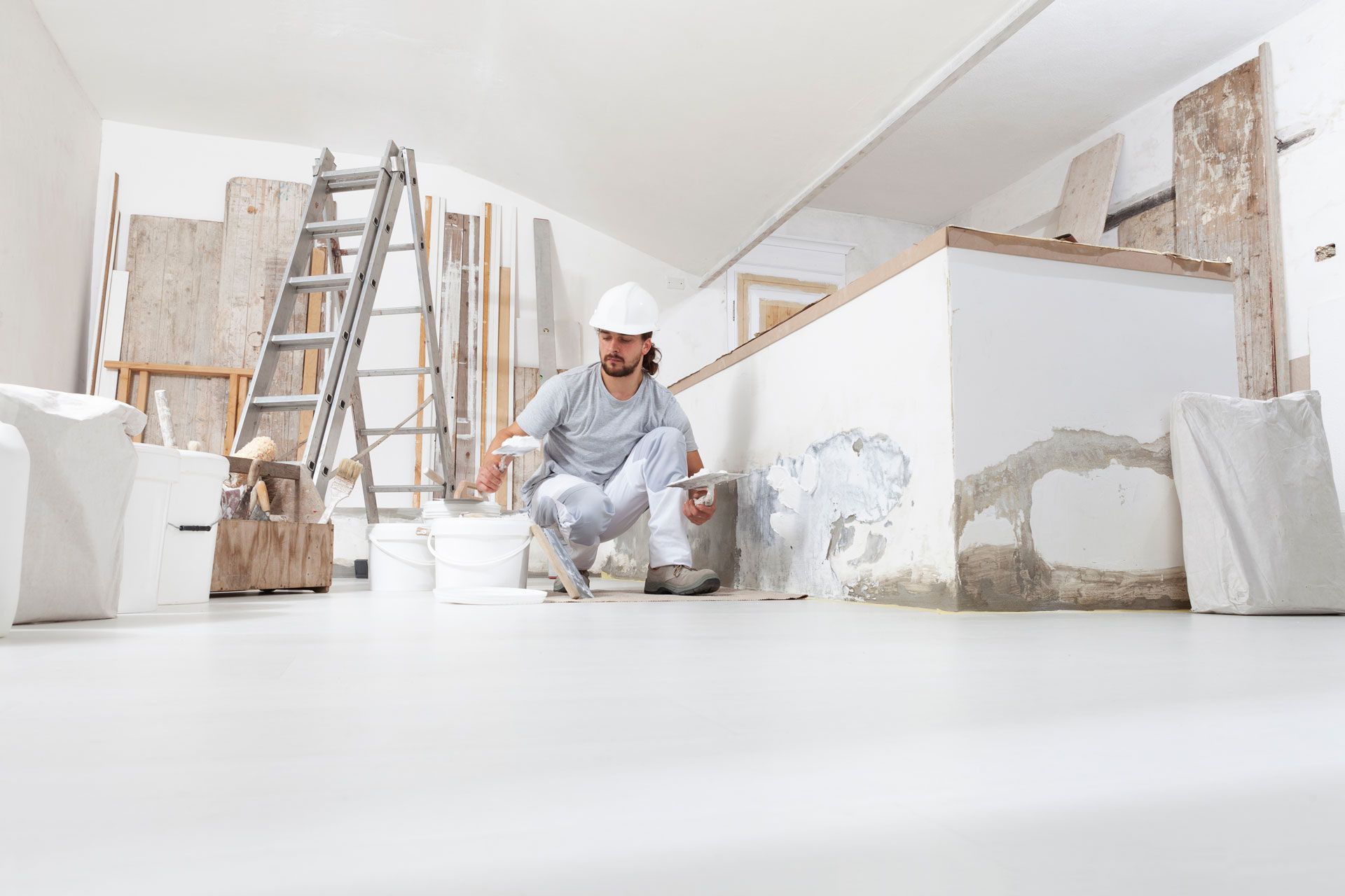 Man in hard hat patching wall damage in a room under renovation, with ladder, buckets, and materials.