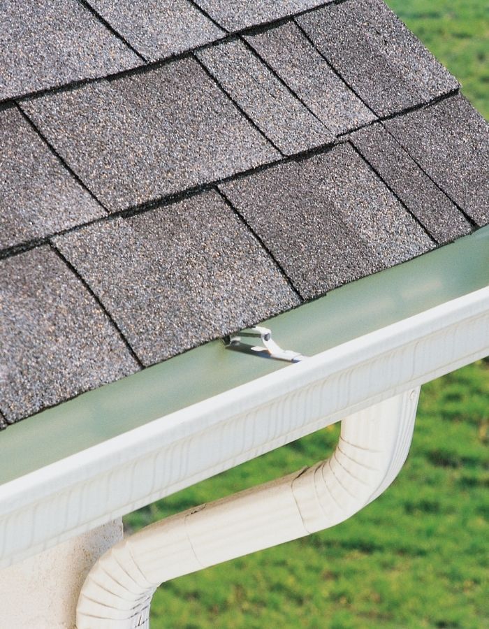 Roof with gray shingles, white gutter and downspout against green grass.