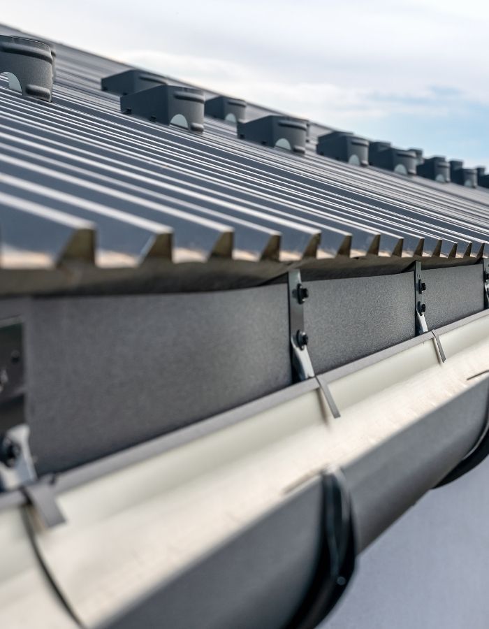 Close-up of a gray metal roof with ridge vents, attached to gray gutters with support brackets.