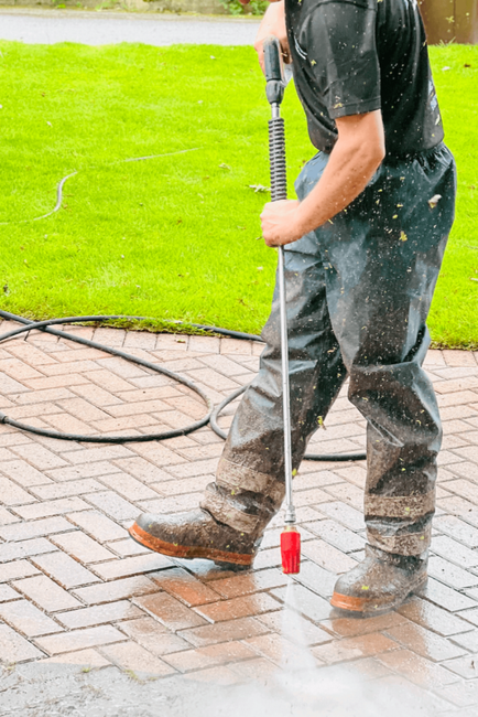 Man holding a pressure washer, smiling, standing on a driveway in front of a house. He's wearing a cap and t-shirt with logo.