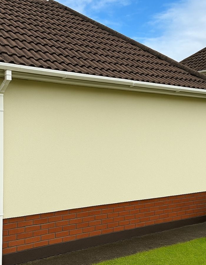 Tan stucco wall with brown brick base, brown tiled roof, and white gutter against a blue sky.