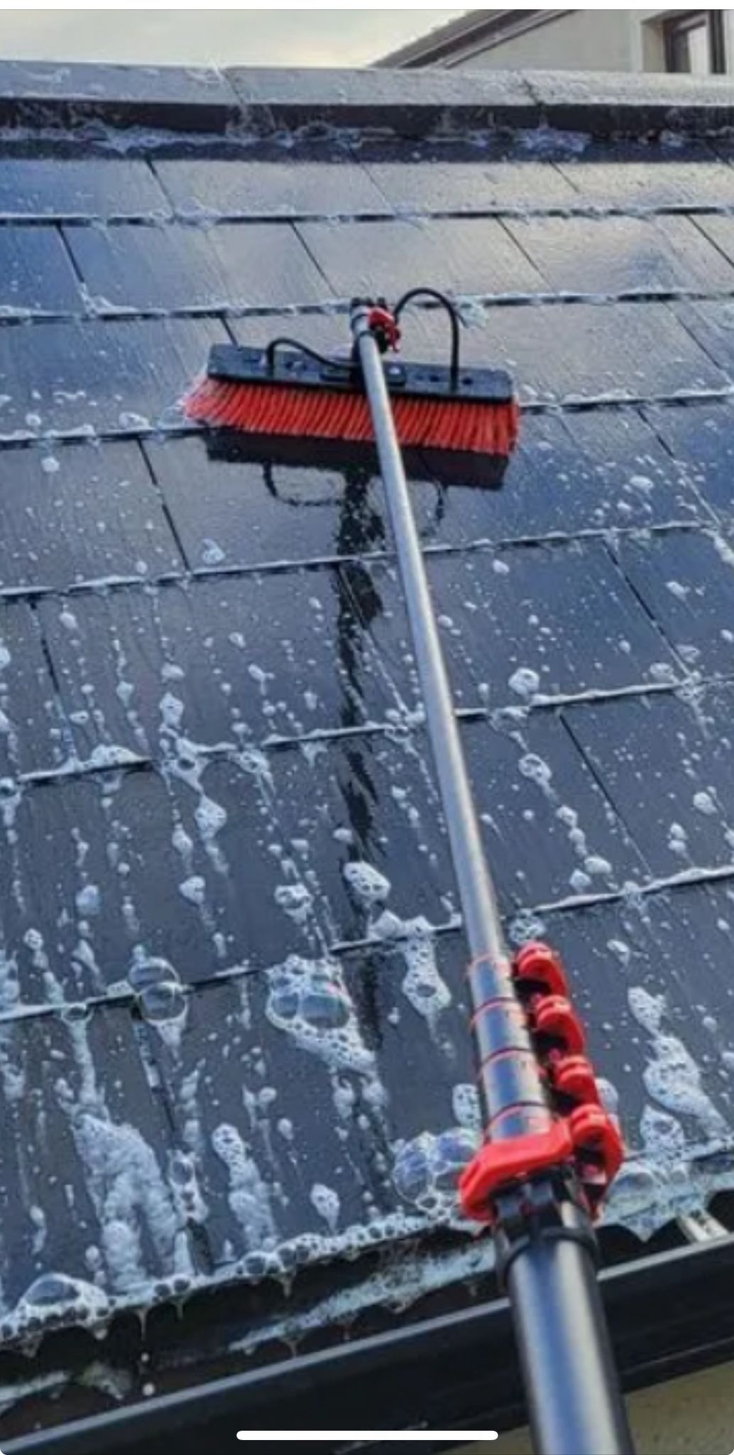 Man on a ladder power washing a roof, removing green moss to reveal red tiles. House exterior.