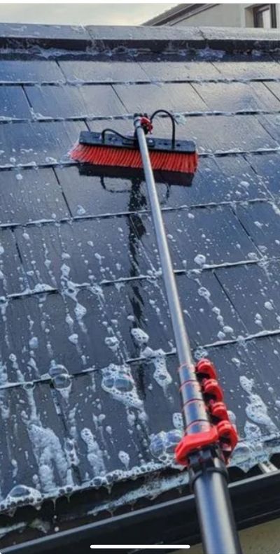 Man on ladder power washing a mossy roof, cleaning it to reveal orange tiles.