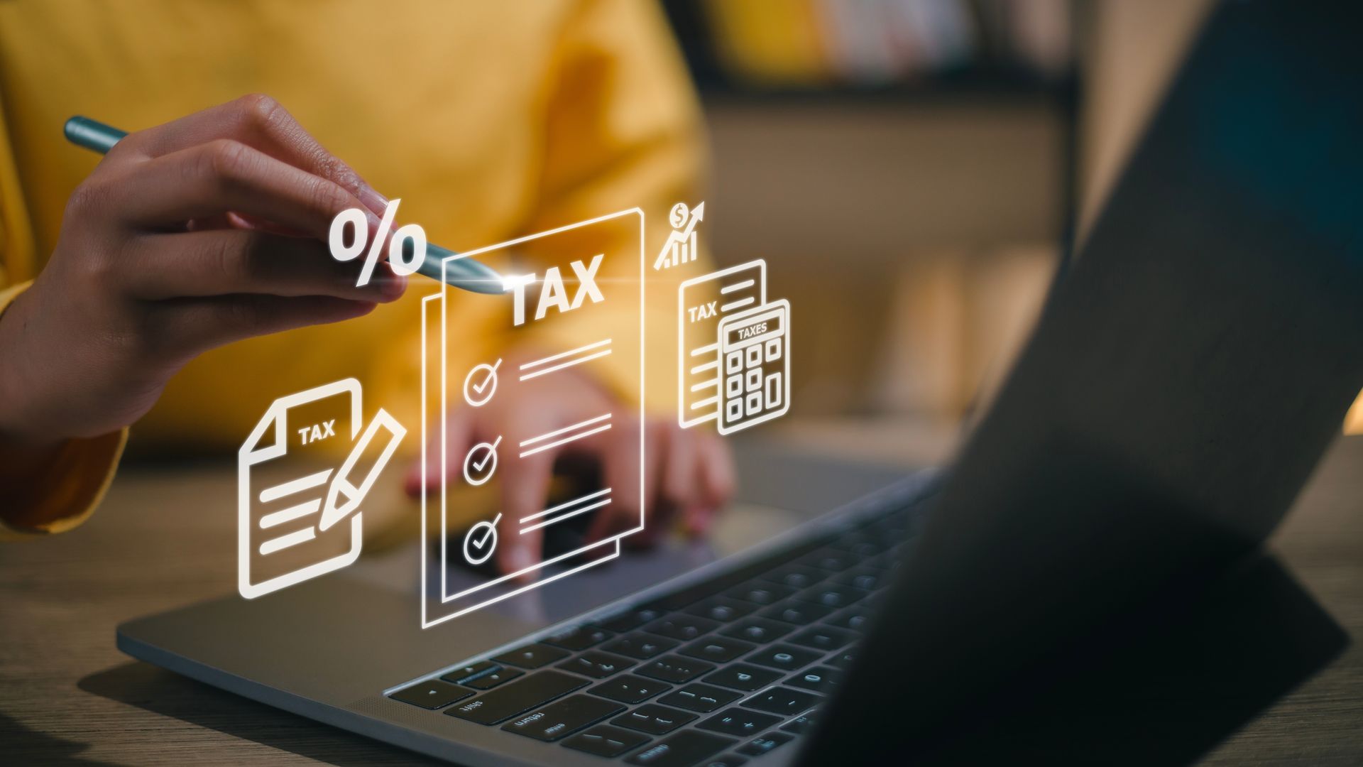 Hand using pen over laptop with holographic tax icons and documents displayed.