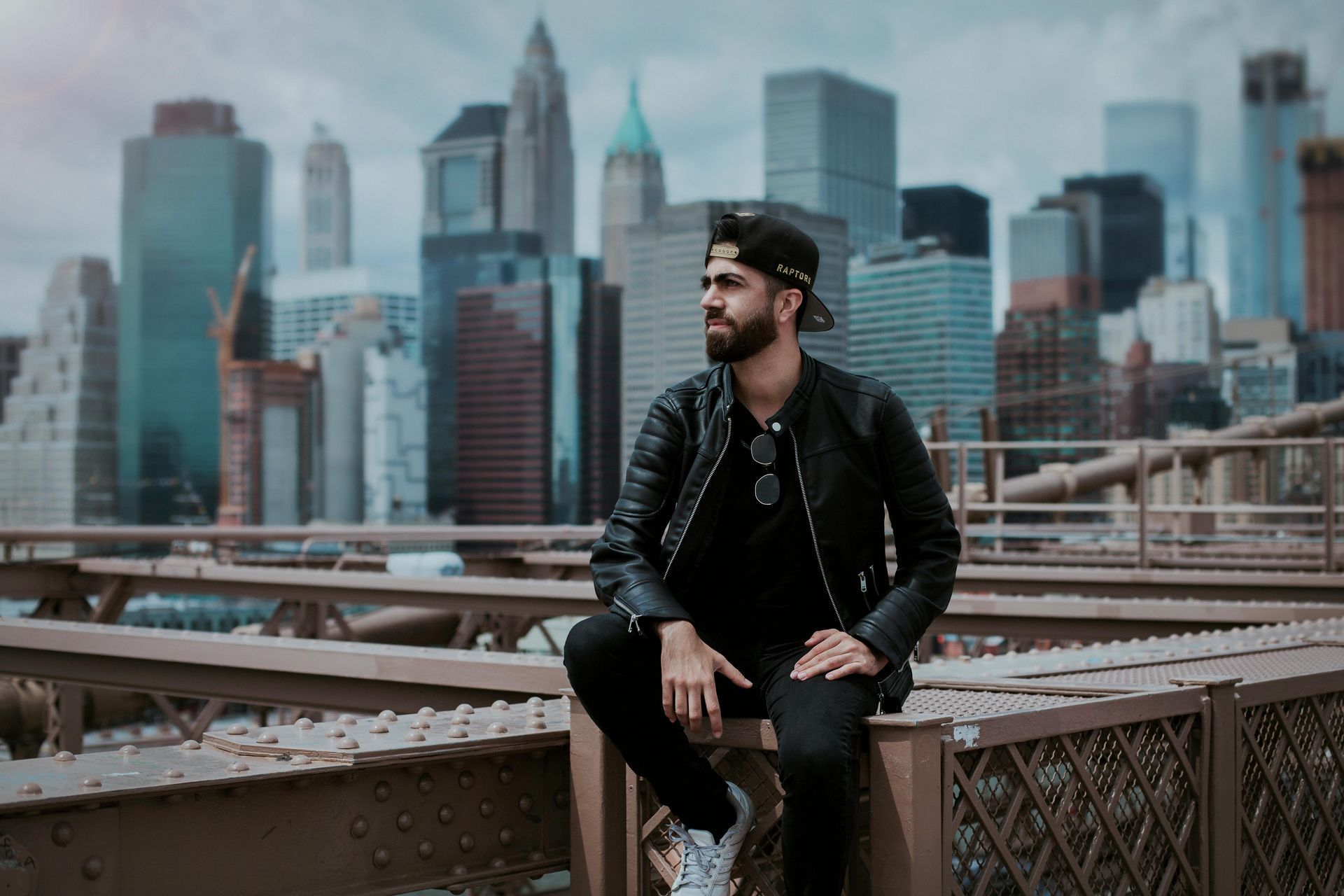 Man wearing a leather jacket sits on a bridge with a city skyline in the background.