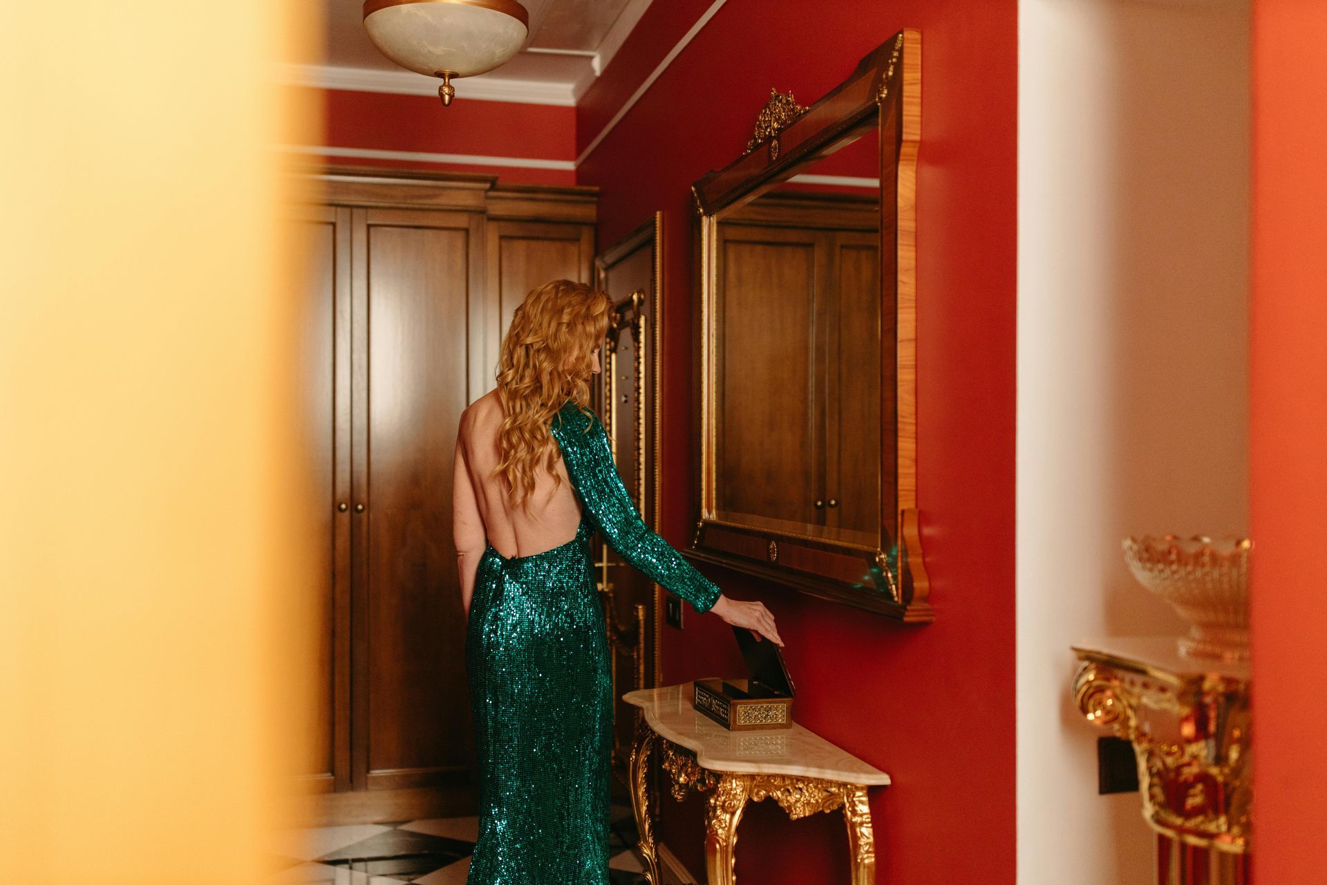 Woman in green sequined gown near ornate mirror and table in a red room.