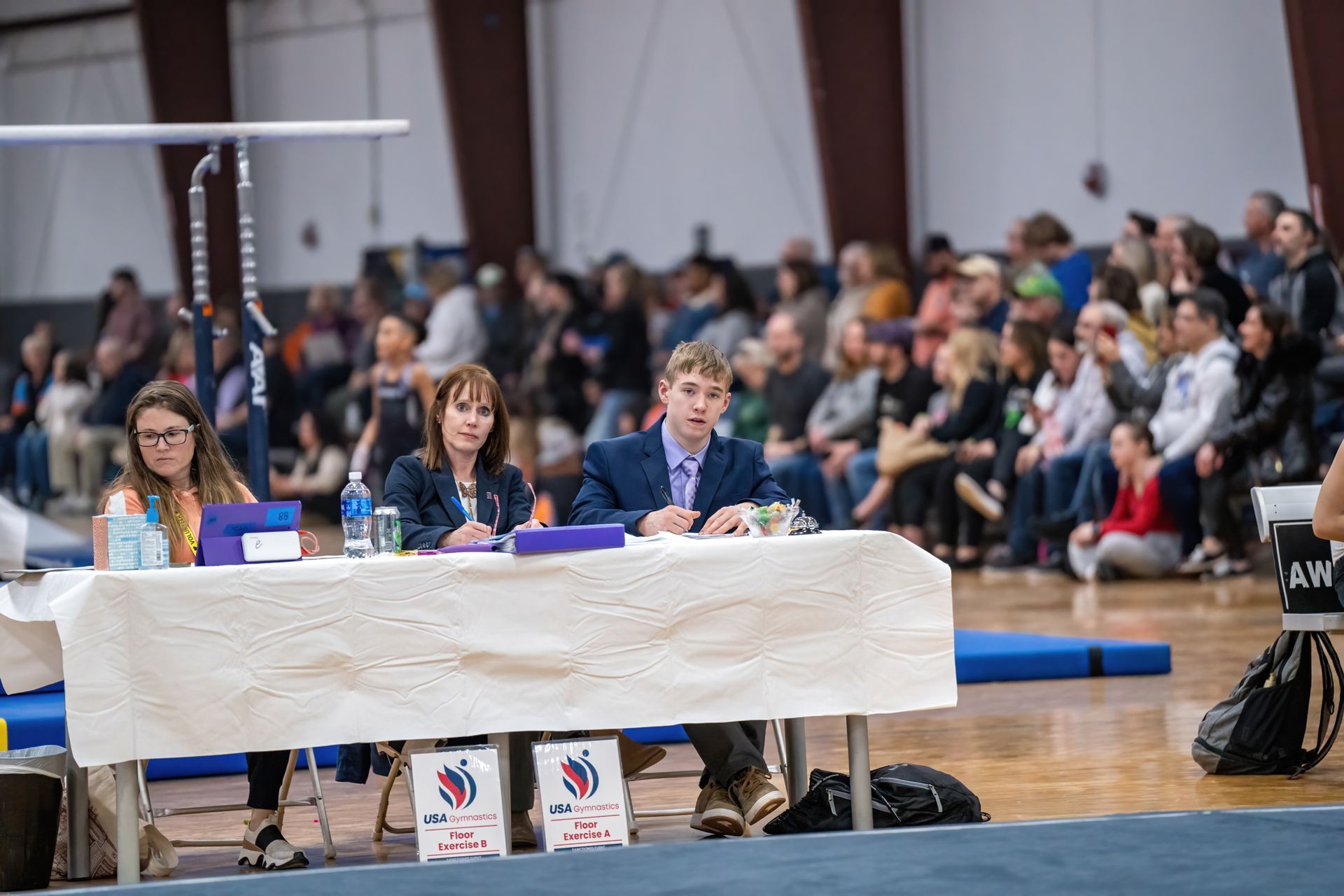 A group of people are sitting at a table in a gym watching a gymnastics competition.