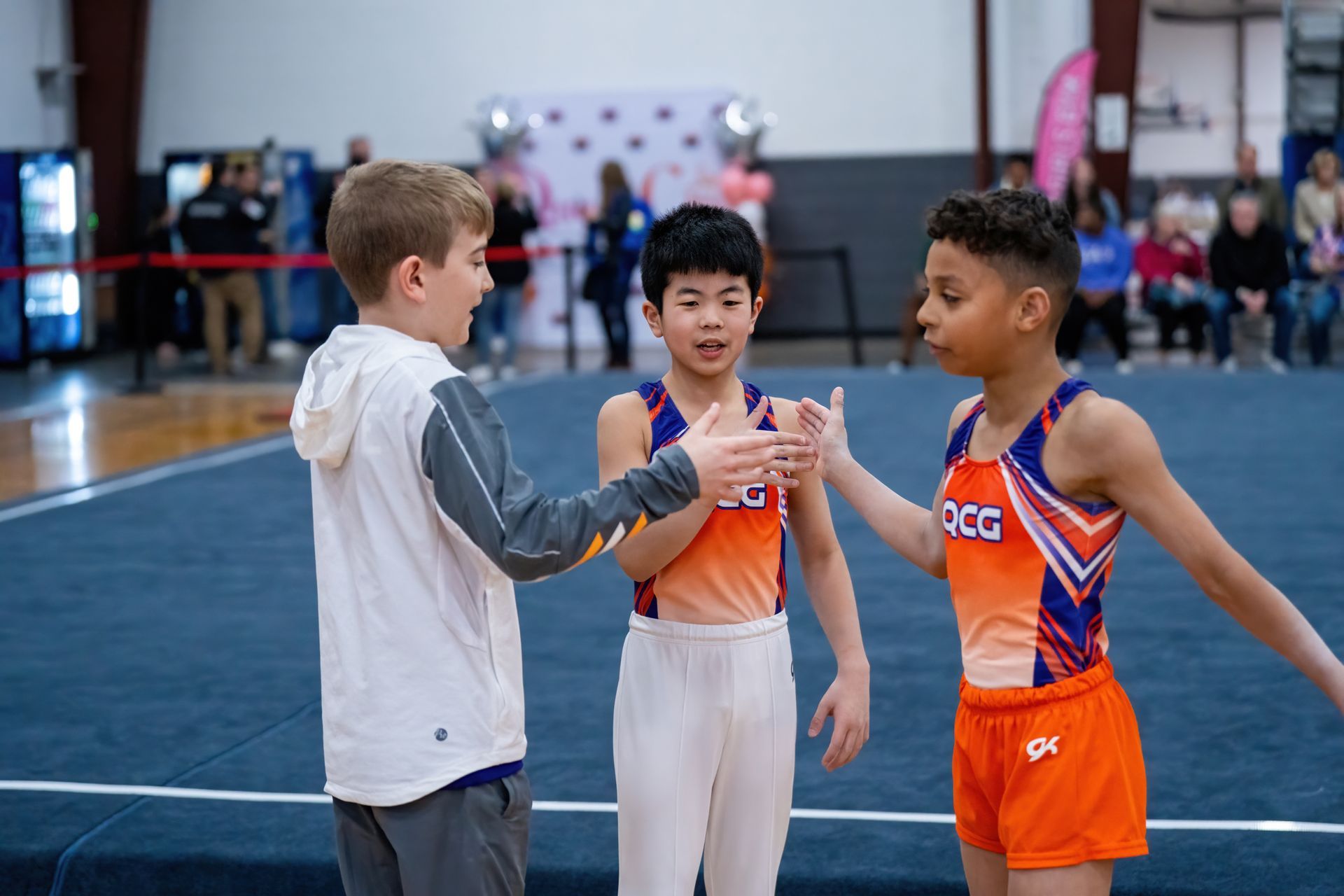 Three young boys are standing on a gymnastics floor talking to each other.