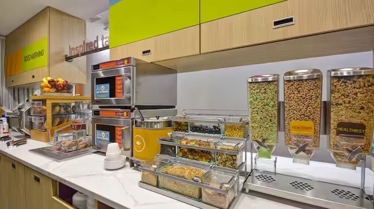 Hotel breakfast bar with cereal dispensers, muffins, pastries, and two stacked microwaves.