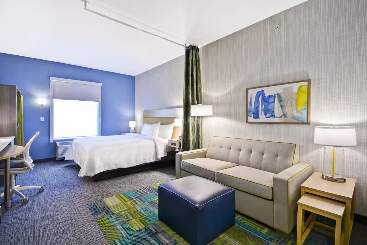 Hotel room with bed, sofa, and desk, blue walls, carpet with green accents, and artwork.