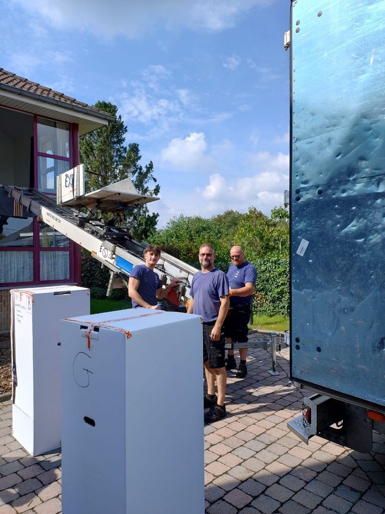 Des cartons empilés dans une pièce lumineuse ; une échelle, du ruban adhésif et des plantes sont également présents, suggérant un déménagement.