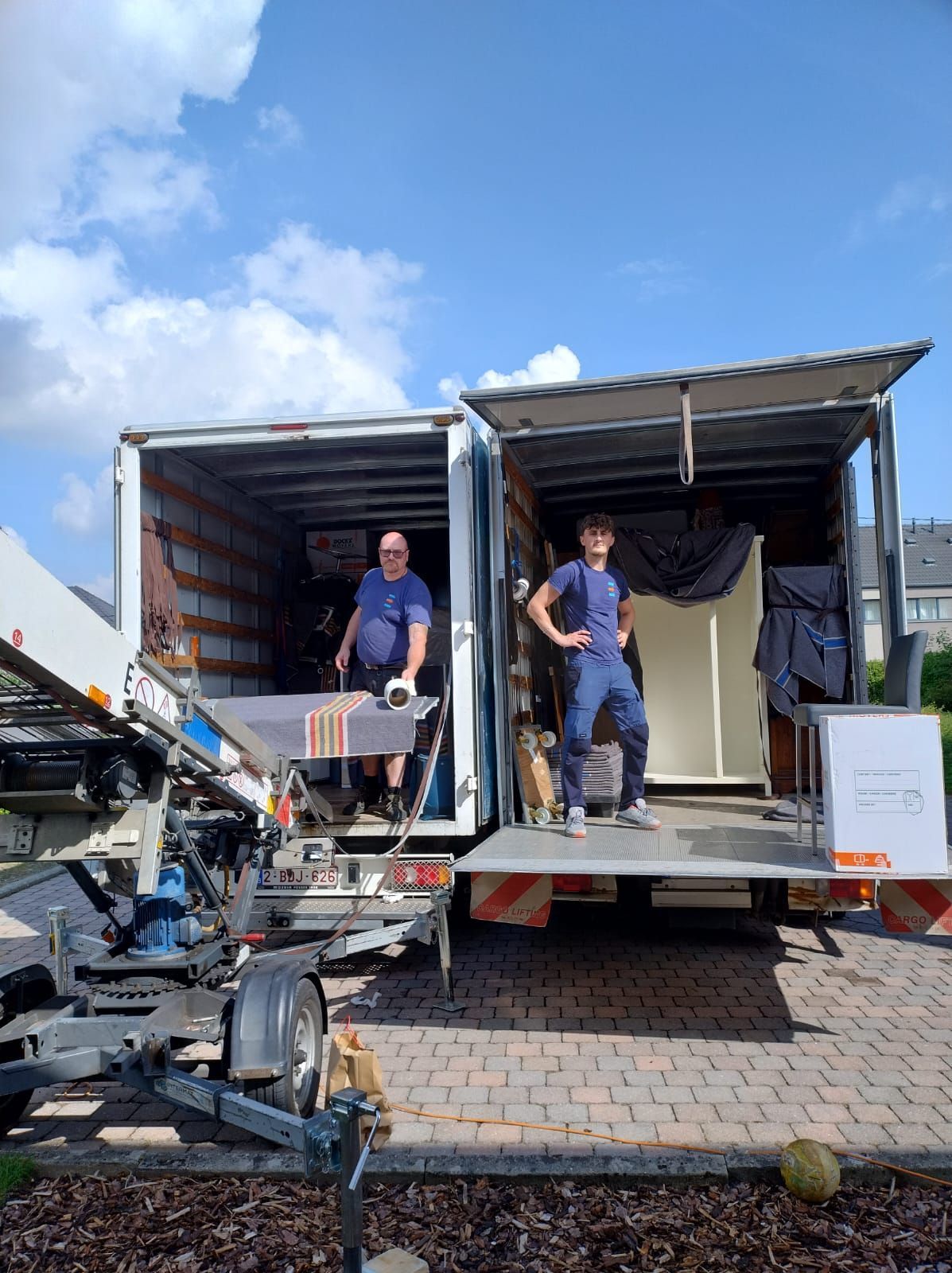 Un homme charge des cartons d'un camion dans une rue de la ville ; une personne passe. Journée ensoleillée.