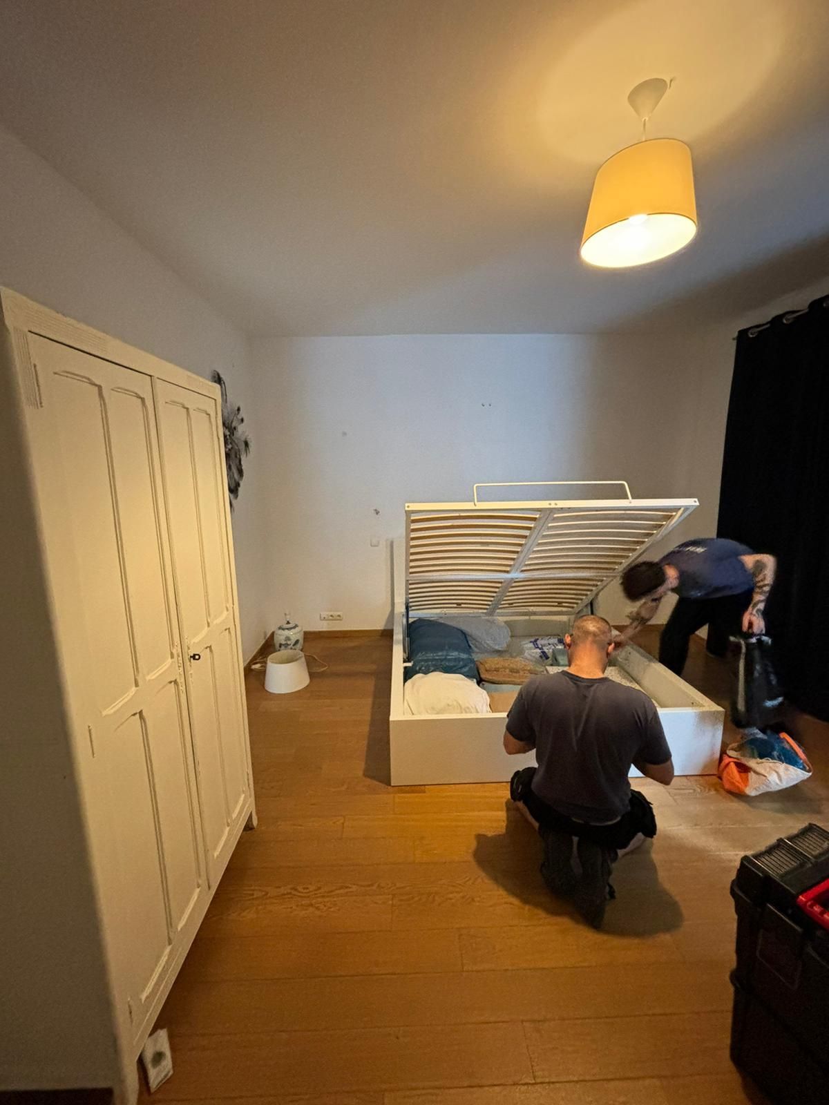 Deux personnes montent un lit dans une chambre avec une armoire blanche et du parquet.