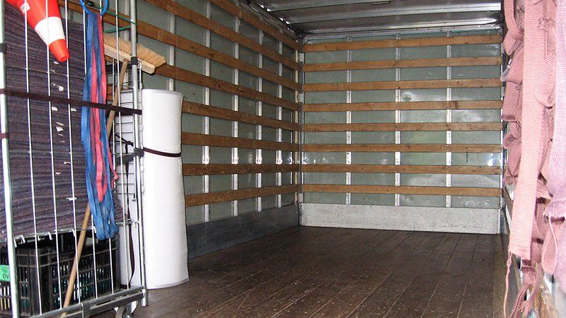 Intérieur d'un camion de marchandises. Plancher et parois marron avec supports en bois. Sangles et autres éléments visibles.