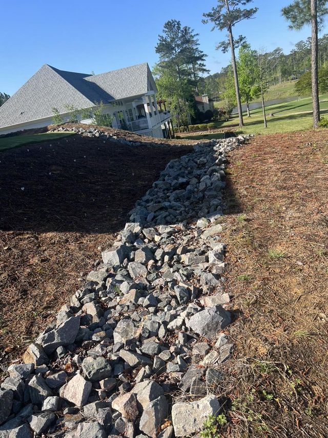 A rock-lined drainage swale runs between a mulch-covered slope and a grassy hillside near a suburban house.
