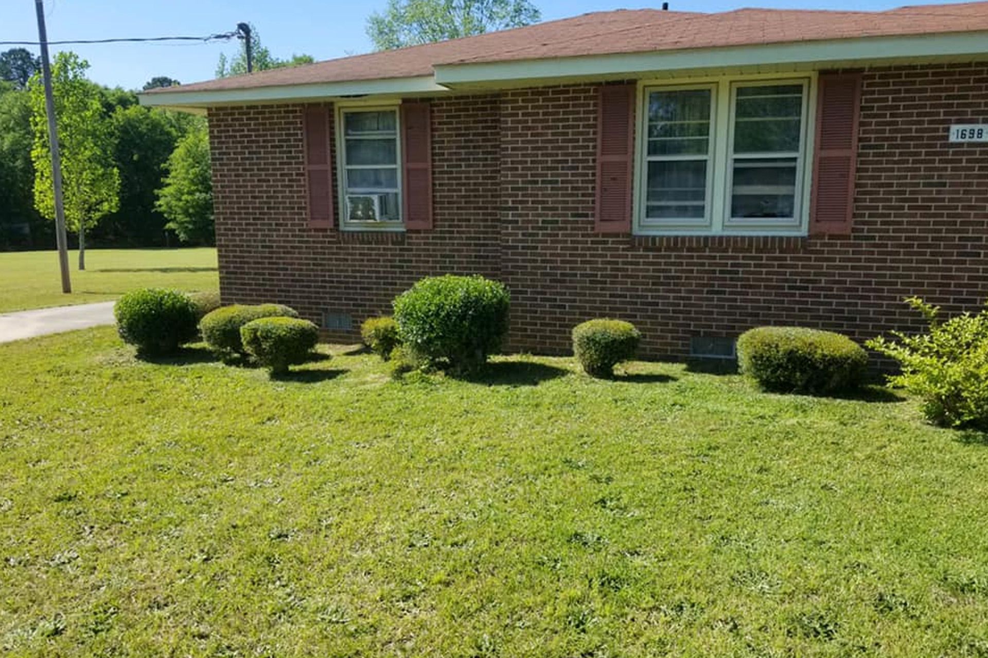 A brown brick, one-story house with a green lawn and small, round bushes in front on a sunny day.