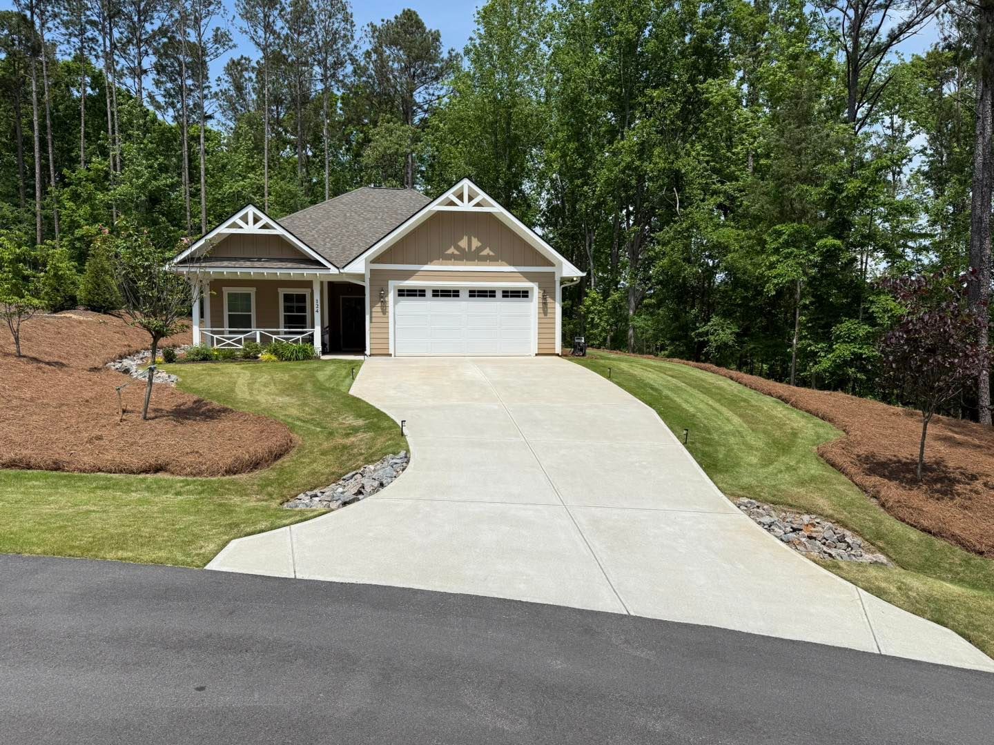 A tan, single-story house with a white garage door and a concrete driveway, surrounded by trees and landscaped mulch beds.