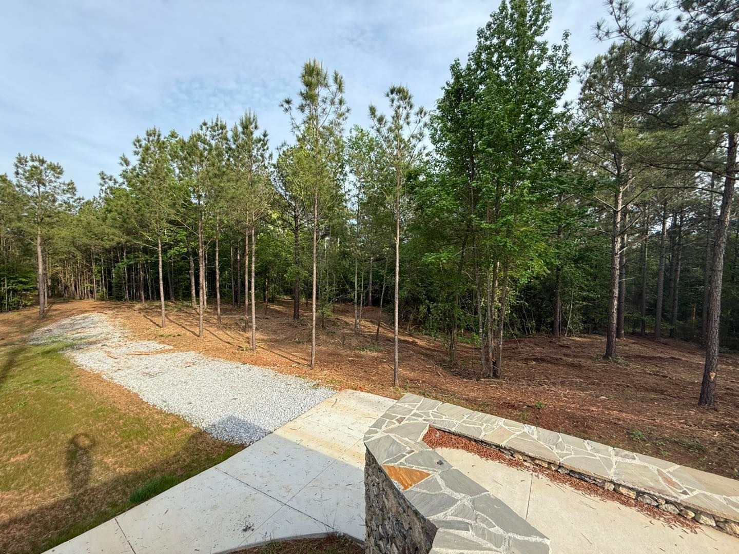 A stone-walled patio leads to a gravel path that winds through a wooded backyard on a sunny day.