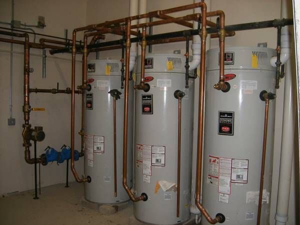 Three grey industrial water heaters connected by copper plumbing against a white wall in a utility room.