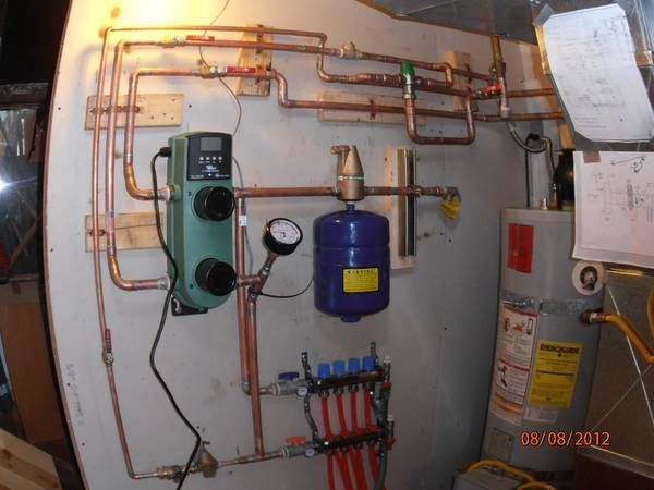 A boiler system with copper piping, a green circulation pump, a blue expansion tank, and a red manifold on a wall.
