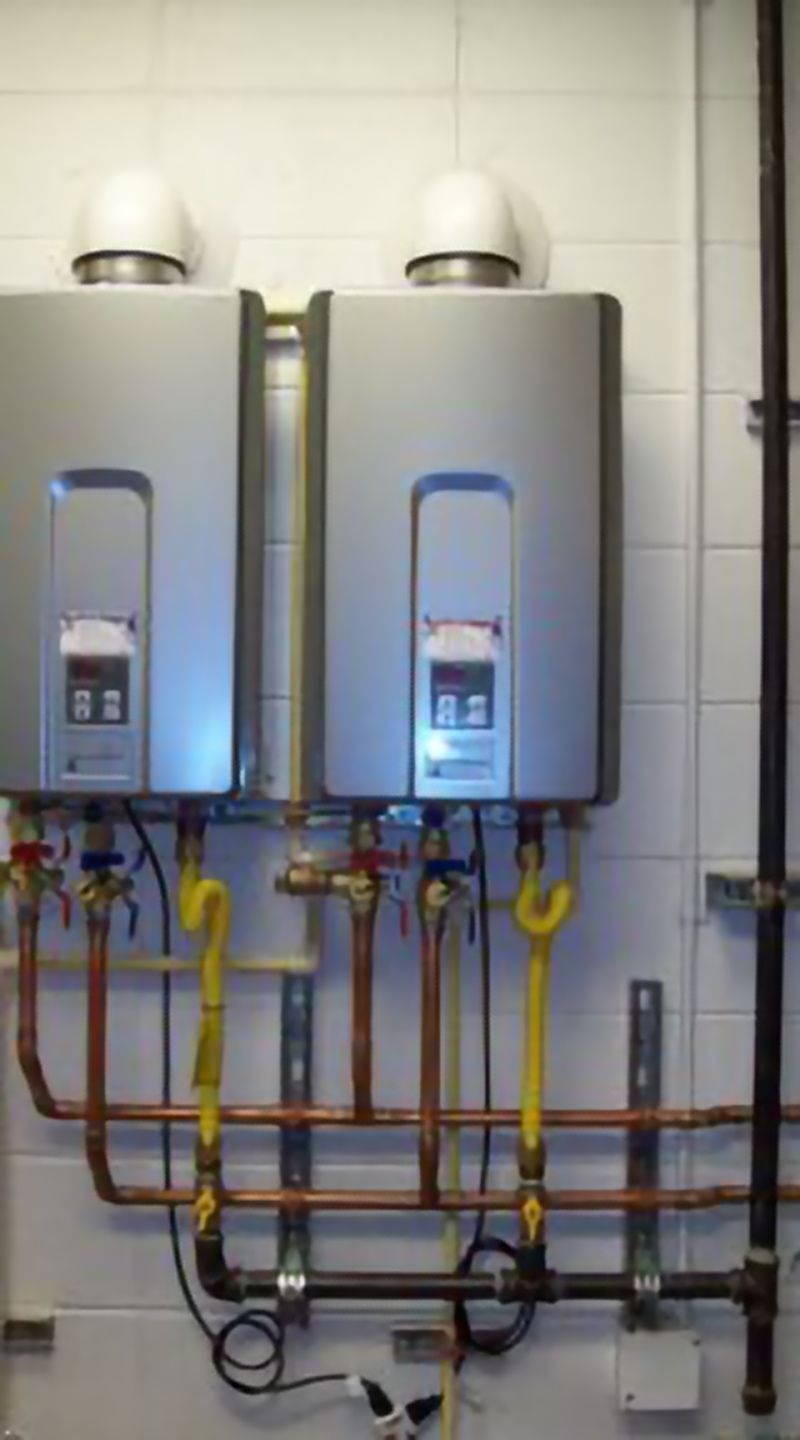 Two silver tankless water heaters mounted on a white brick wall with connected copper pipes and yellow gas lines.