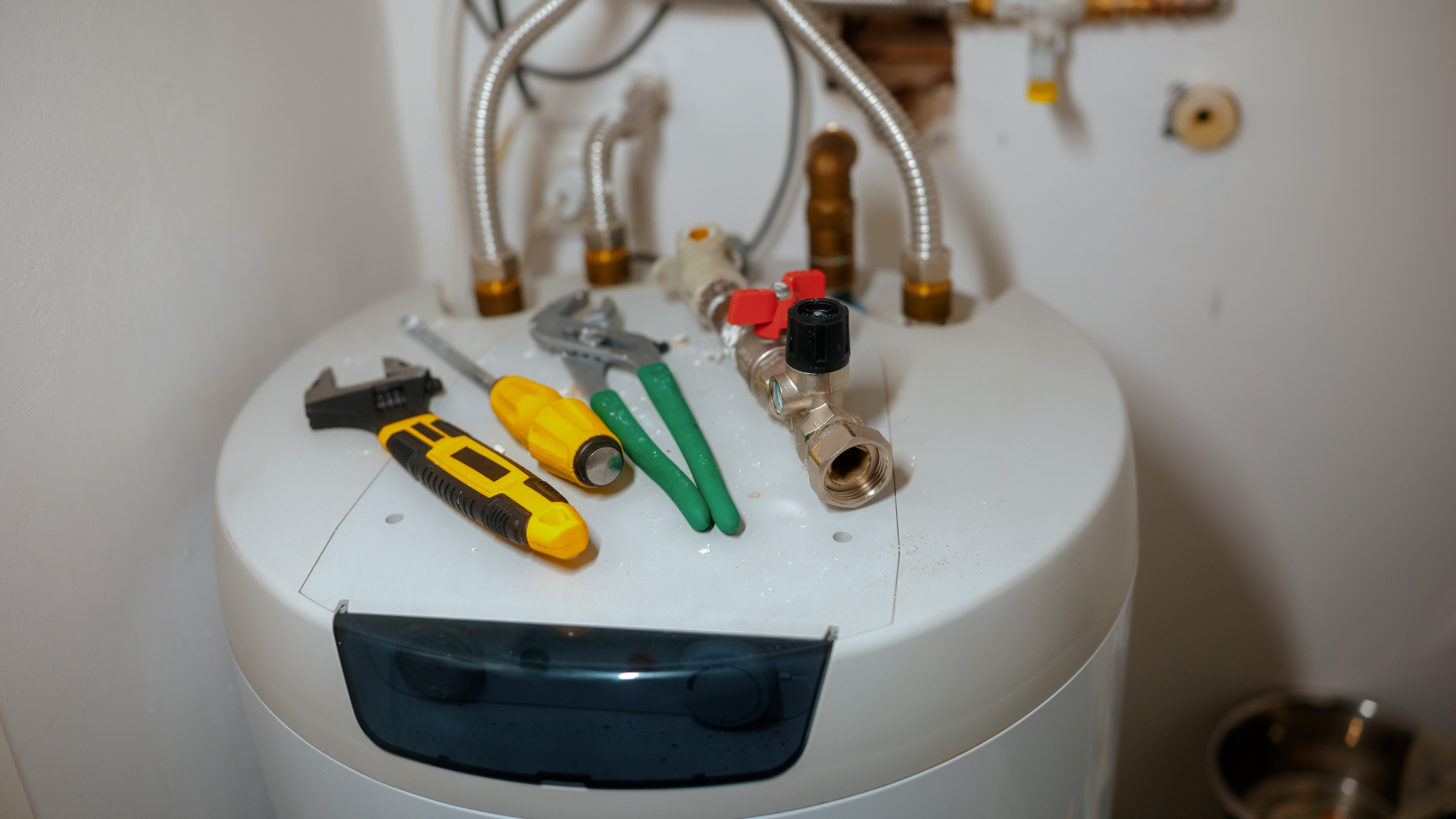 Tools, including a wrench, screwdriver, and pliers, resting on top of a water heater with plumbing components.