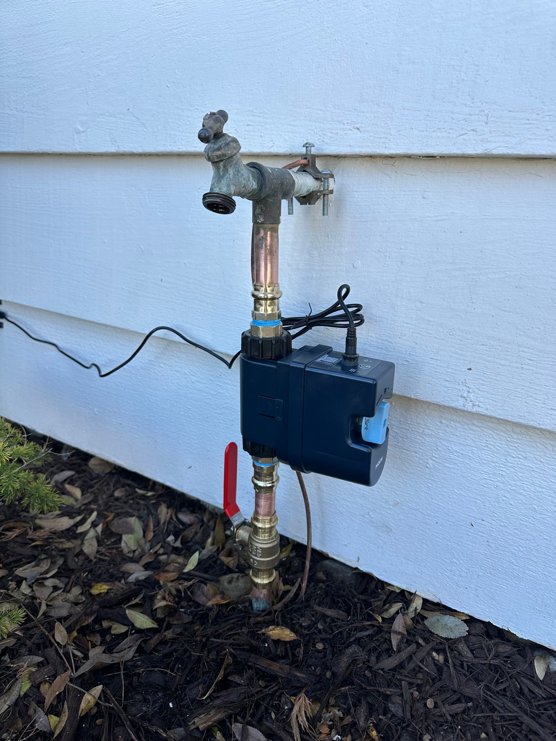 Outdoor water spigot with attached irrigation valve and wiring. Brown mulch in front.