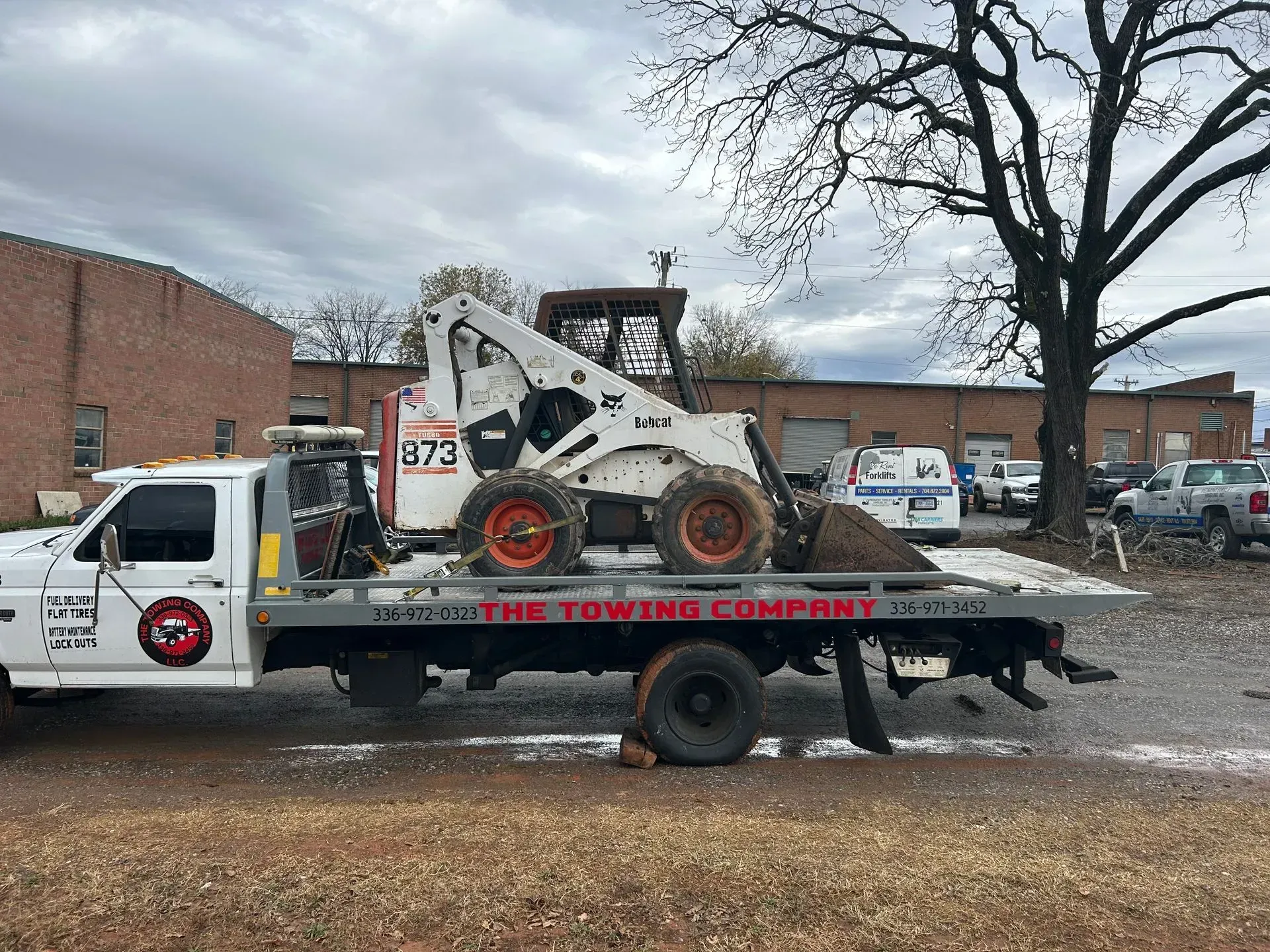 Tow truck carrying a white skid steer loader on a flatbed in front of brick buildings and bare trees.