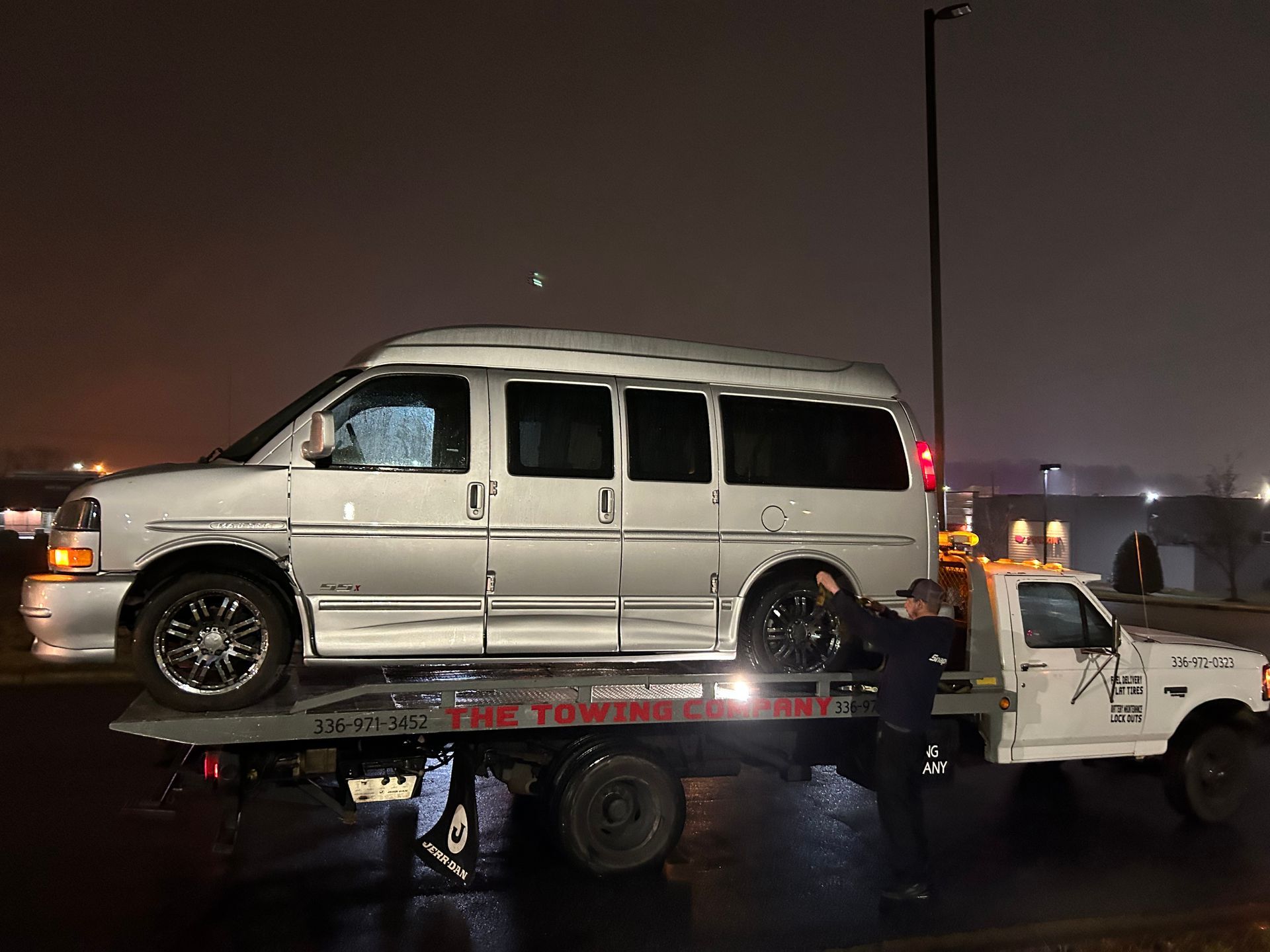 A white van is being towed by a tow truck at night.