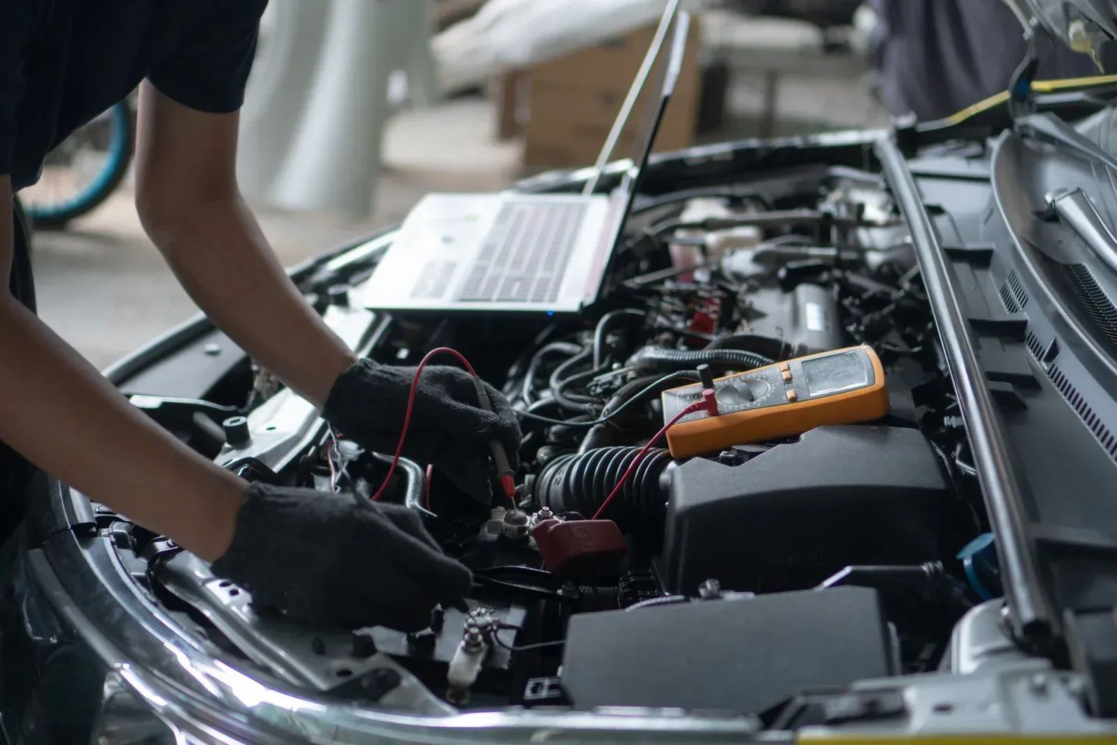 Mechanic using a laptop and multimeter to diagnose a car engine. Black gloves, open hood, indoor setting.