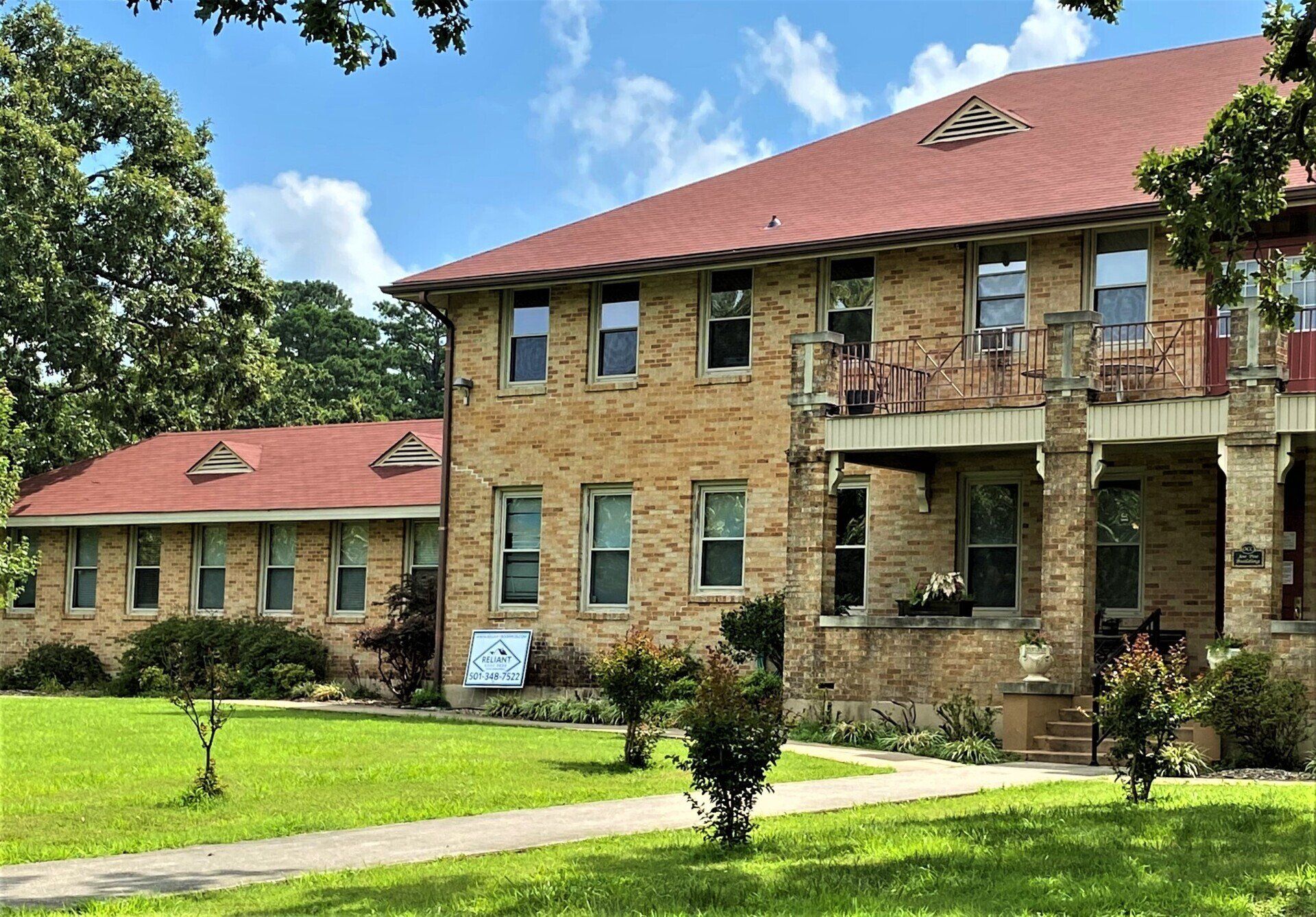 Two-story brick building with red roof, green lawn, and trees under a partly cloudy sky.