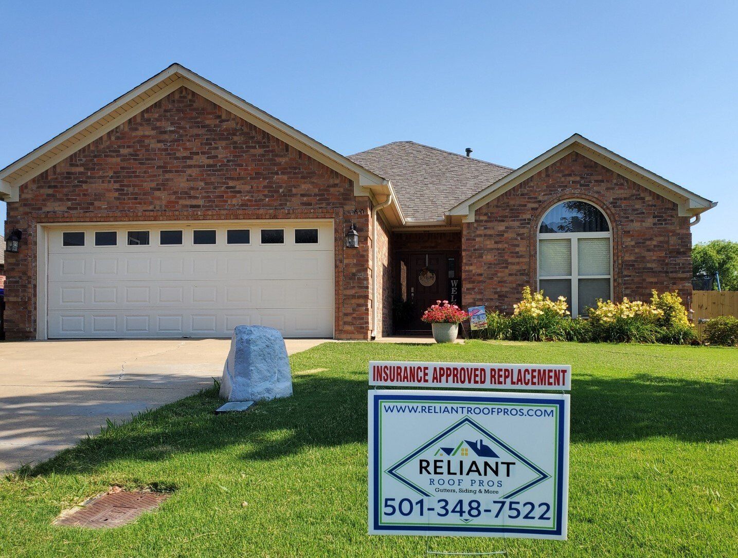 A brick house with a white garage door and a reliant roofing sign in front of it.