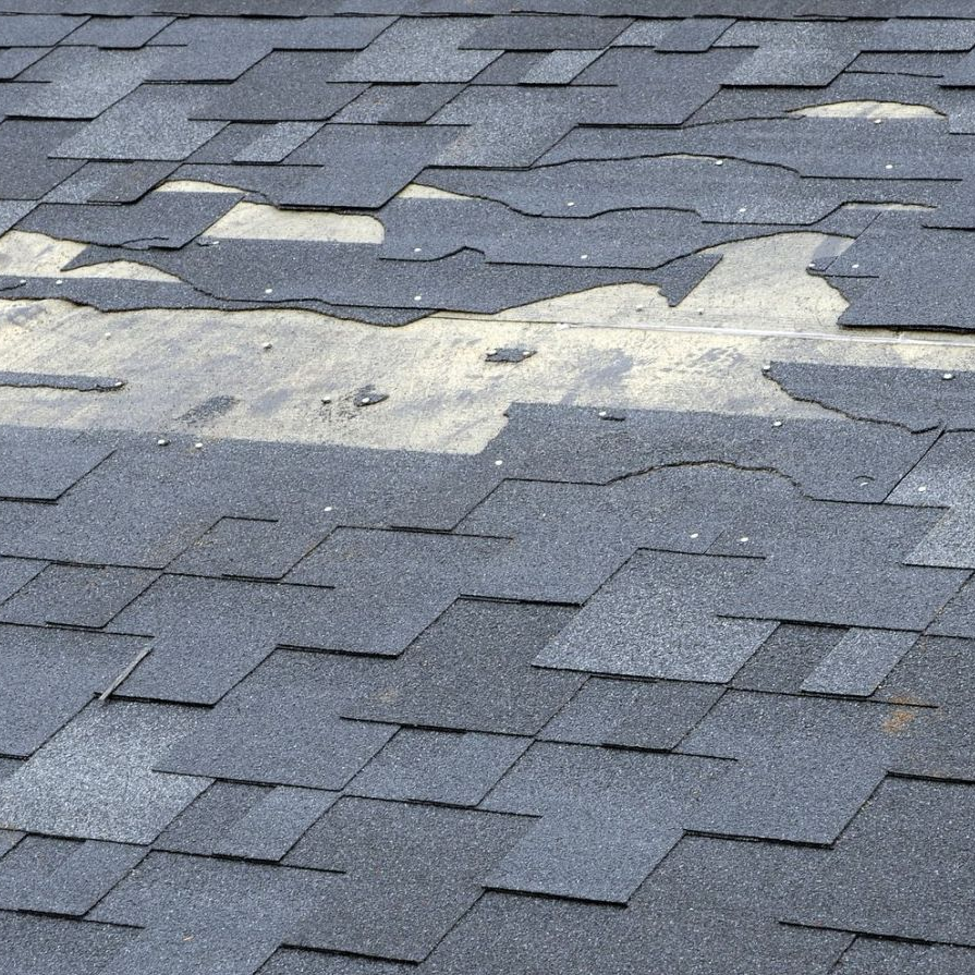 Damaged gray roof shingles with a large patch of missing material exposing lighter underlayment