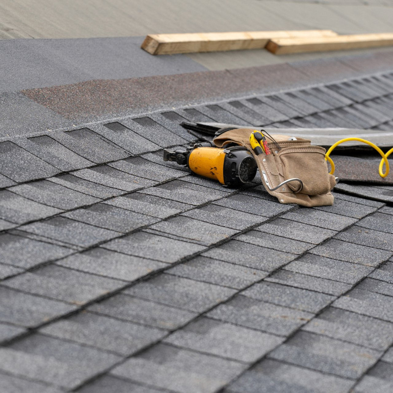 Worker using a yellow nail gun to install gray shingles on a roof